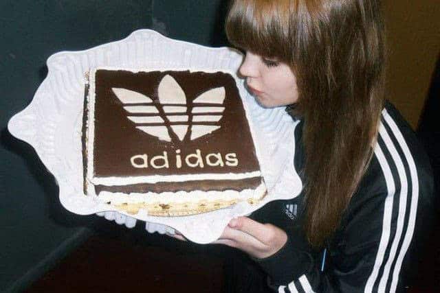 A young woman wearing a classic black three-stripe Adidas tracksuit leaning over a custom-made square chocolate cake featuring the Adidas trefoil logo.