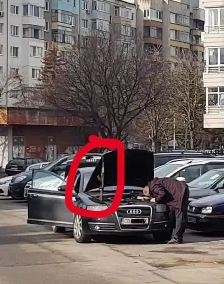A funny Russia picture showing a man inspecting the engine of an Audi in a parking lot, using a long-barrel firearm as a makeshift prop to hold the car's hood open.