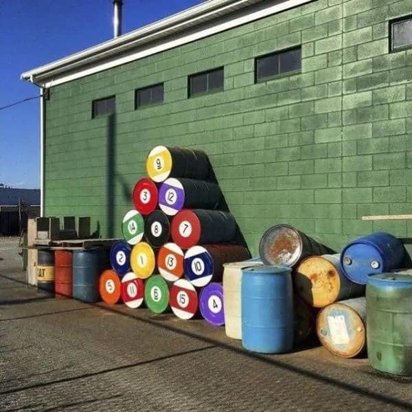 A satisfying visual captured in a trending meme dump features a large stack of industrial oil drums precisely painted and numbered to look like a full set of billiard pool balls arranged in a perfect triangle against a green concrete block wall.