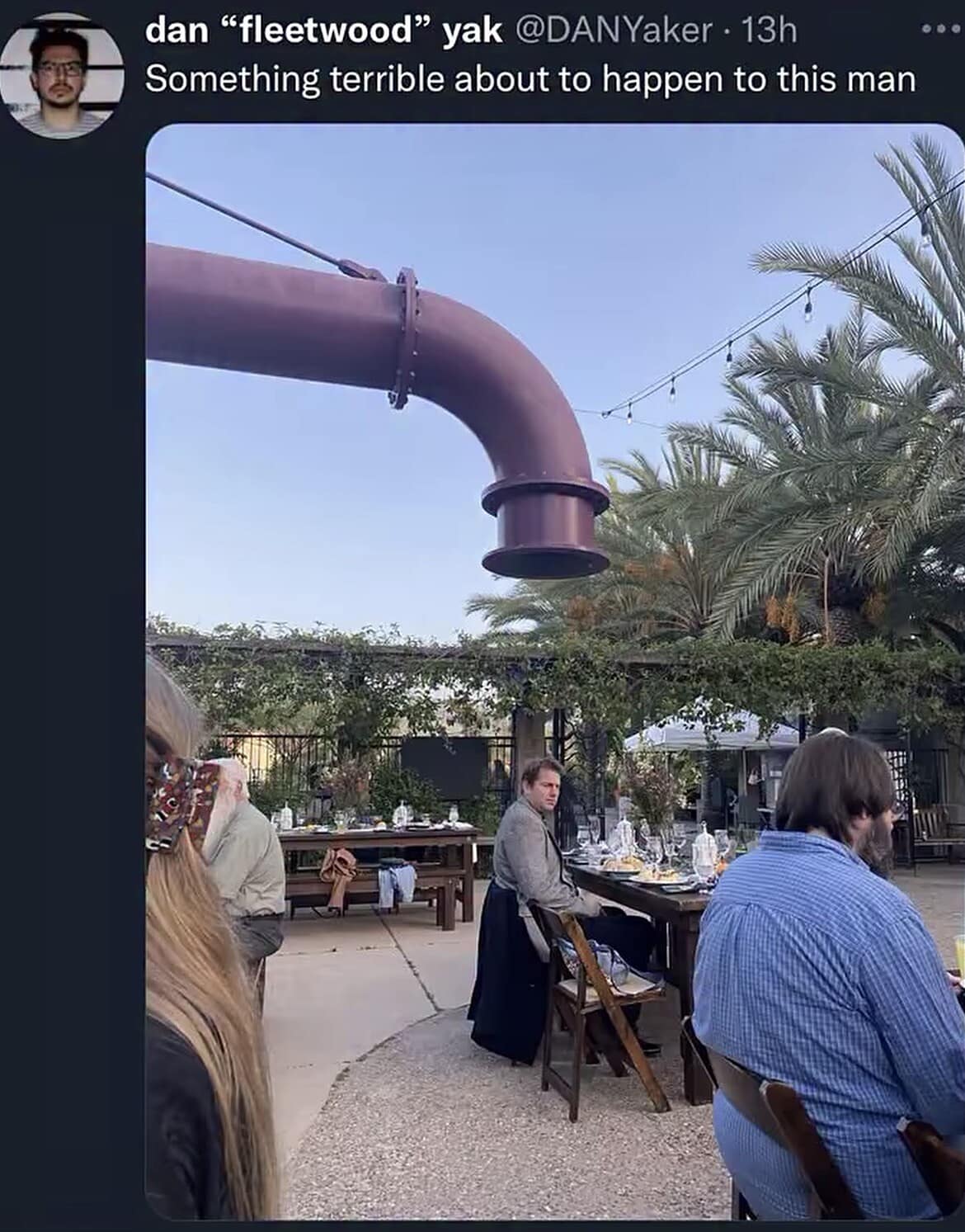 An ominous outdoor restaurant patio photo shows an enormous, curved purple industrial plumbing pipe hanging directly over the head of an unsuspecting lone diner.