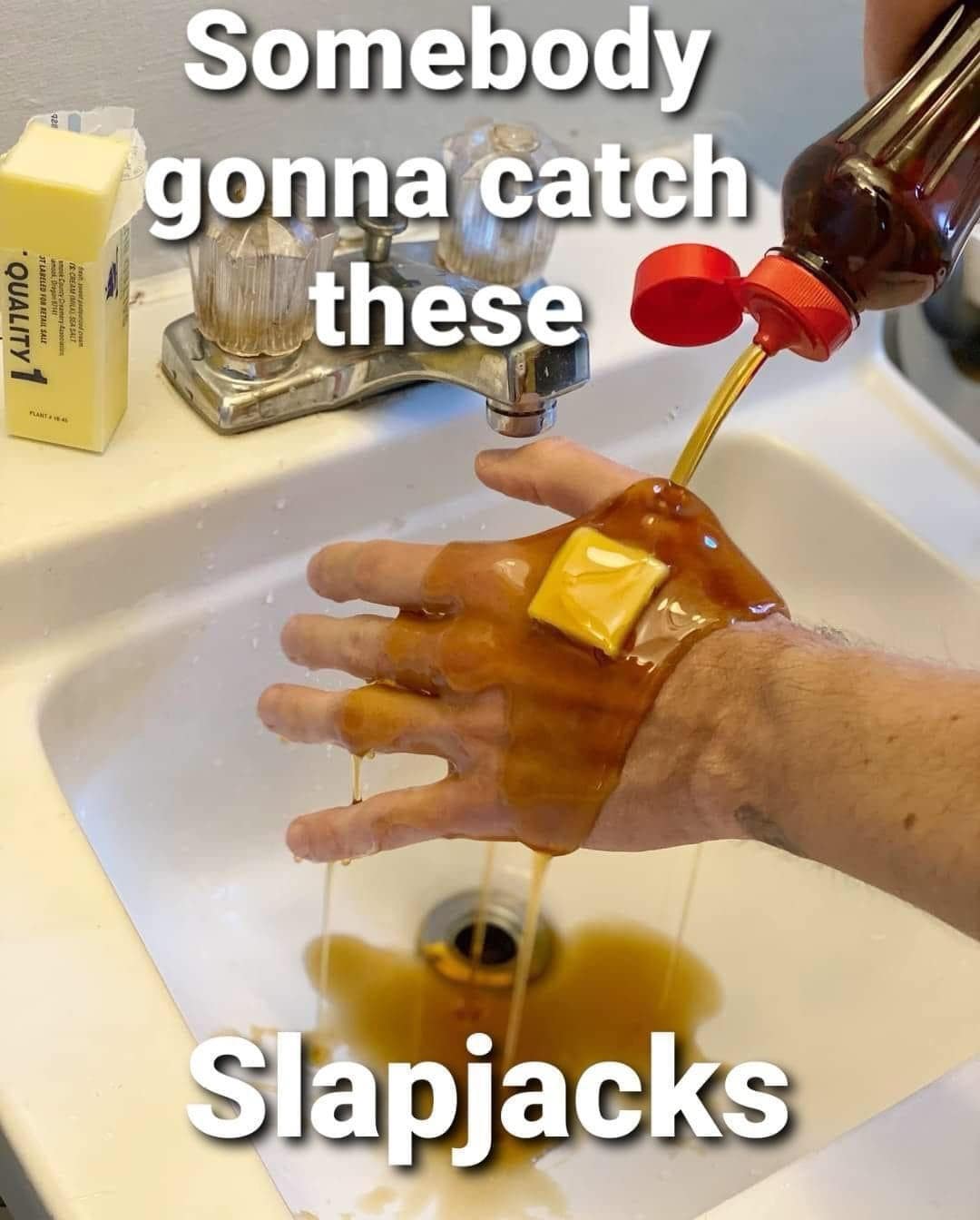 A person pours thick maple syrup onto a pat of yellow butter resting on the back of their hand over a bathroom sink, creating a literal pun for a funny meme about catching hands or "slapjacks."