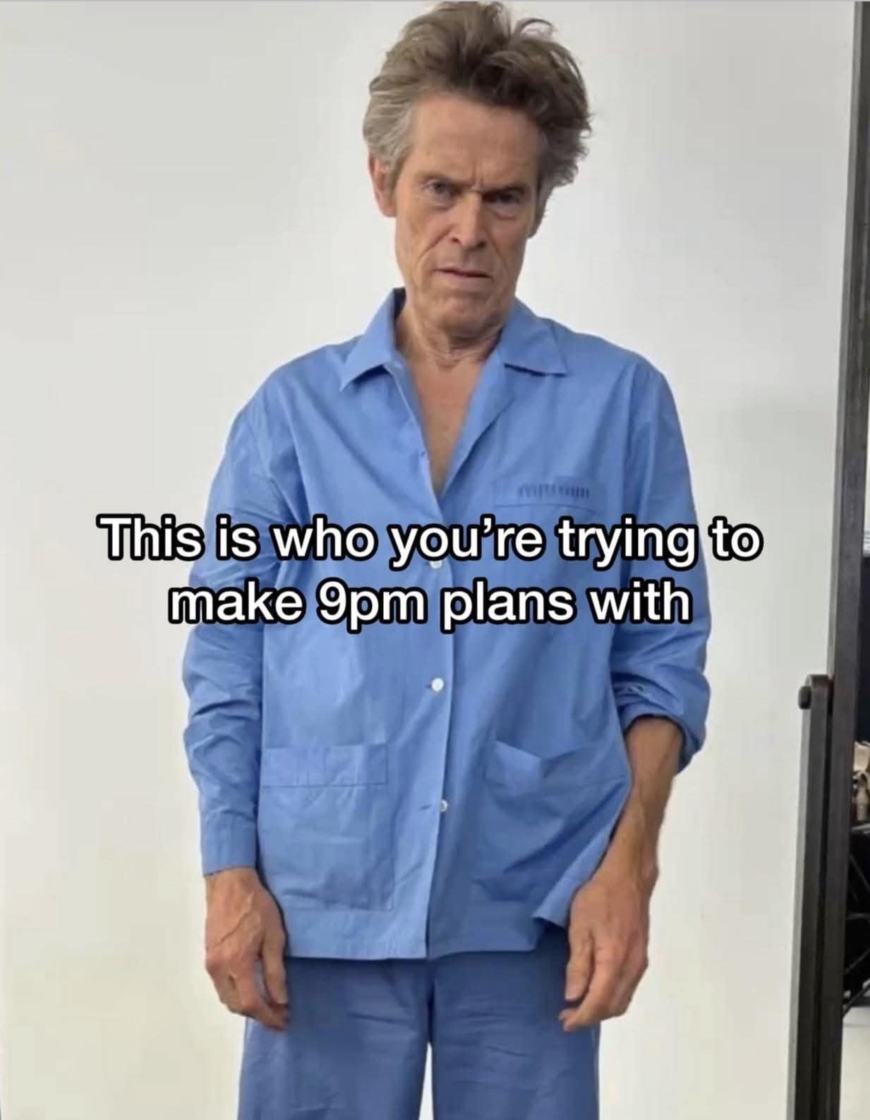 A relatable lifestyle macro features actor Willem Dafoe looking completely exhausted, disheveled, and unblinking in a loose light-blue shirt, captioned perfectly as the physical embodiment of who you are trying to make 9:00 PM plans with.