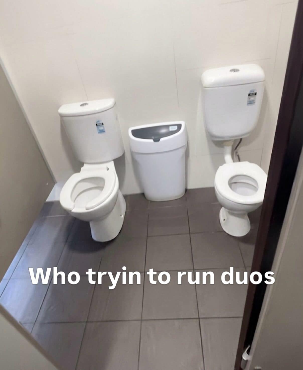 A chaotic hilarious meme showing a public bathroom where two toilets are installed side-by-side with no stall or divider between them, captioned with the gaming phrase: "Who tryin to run duos."