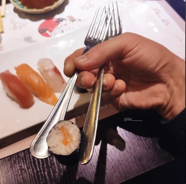 culinary fail photo showing a hand awkwardly gripping two metal forks like oversized chopsticks to lift a single piece of salmon sushi.