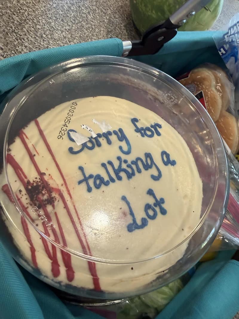 A grocery basket photo shows a white supermarket sheet cake in a plastic container with the message "Sorry for talking a lot" written across the frosting in blue cursive icing.