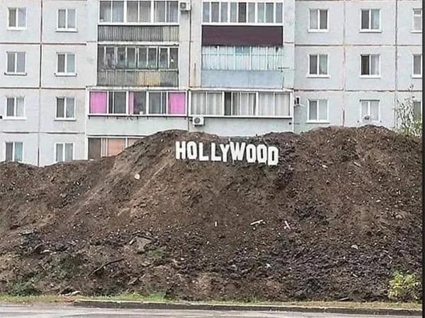 A funny meme showing a depressing, massive mound of brown dirt in front of a gray, Soviet-style apartment building, with the word "HOLLYWOOD" spelled out in small white letters on the slope.