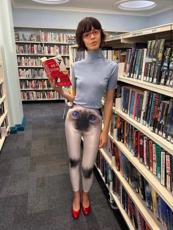 A cursed fashion meme dump photo of a woman in a library wearing white leggings with a massive, high-definition Siamese cat face printed across the entire pelvic region, its blue eyes staring directly at the viewer.