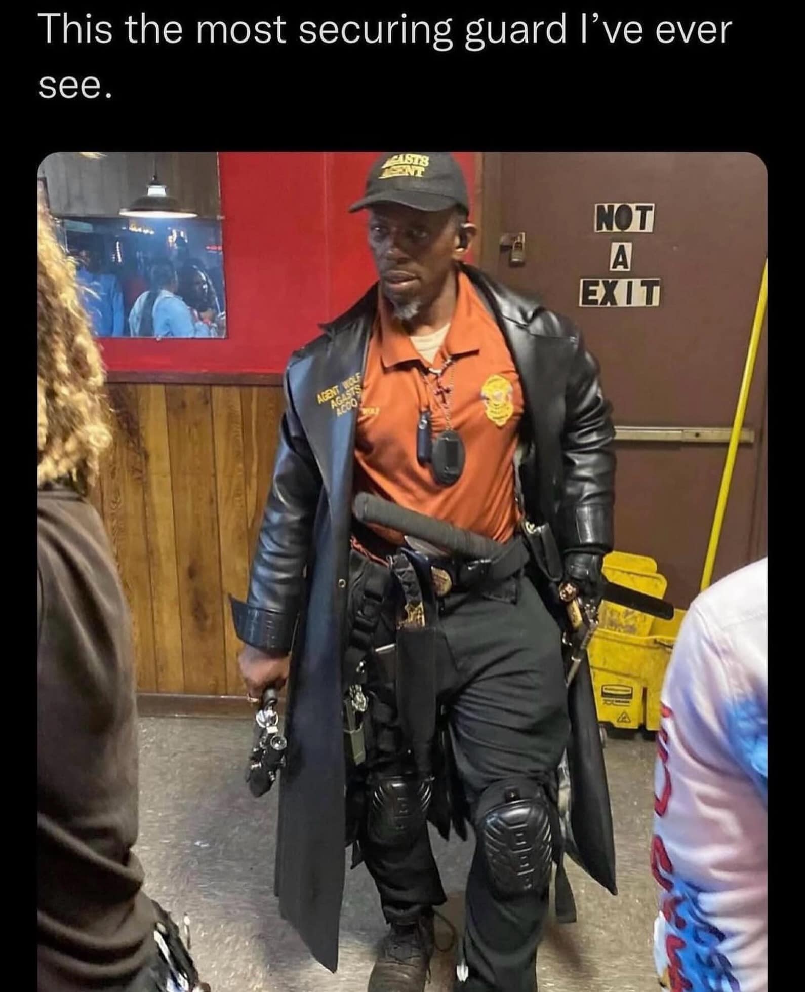 A photo of a mall security guard who has ascended to final-boss status, wearing a full-length black leather duster, tactical knee pads, and carrying an excessive arsenal of knives and gear.