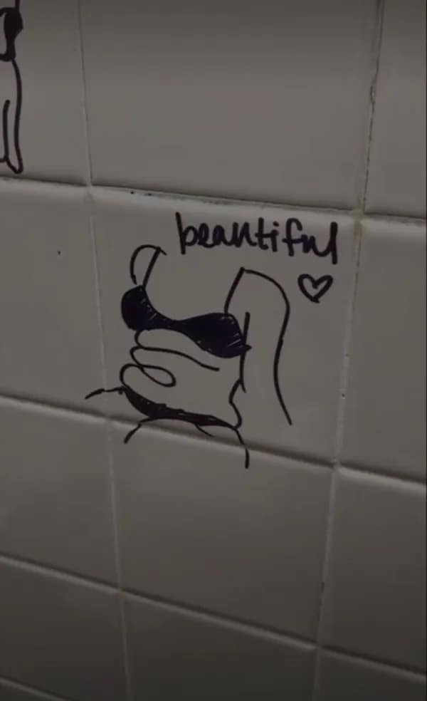 A minimalist black marker drawing on white public restroom tiles showing an illustrative body-positive cartoon figure cleverly sketched around a natural skin crease, labeled with the word "beautiful."