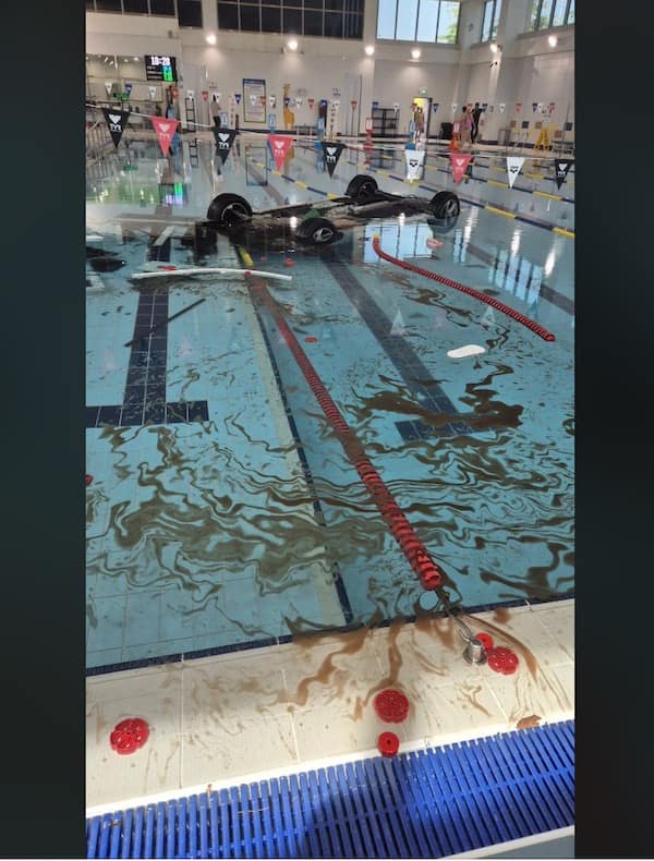 An absolute public facility disaster photo captures an entire car chassis flipped completely upside down and submerged inside a clean, indoor community swimming pool, leaking dark engine oil everywhere.