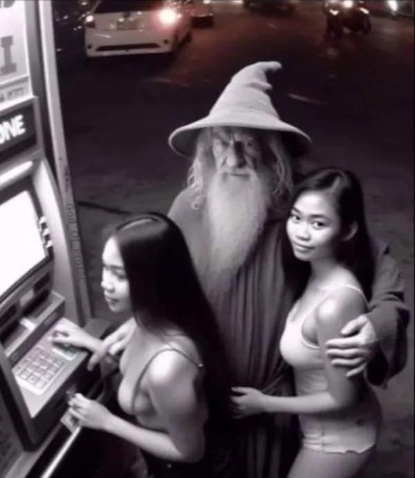 A black-and-white night-vision surveillance style snapshot showing a man fully dressed as the wizard Gandalf from Lord of the Rings with his arms wrapped around two women using an outdoor street ATM.