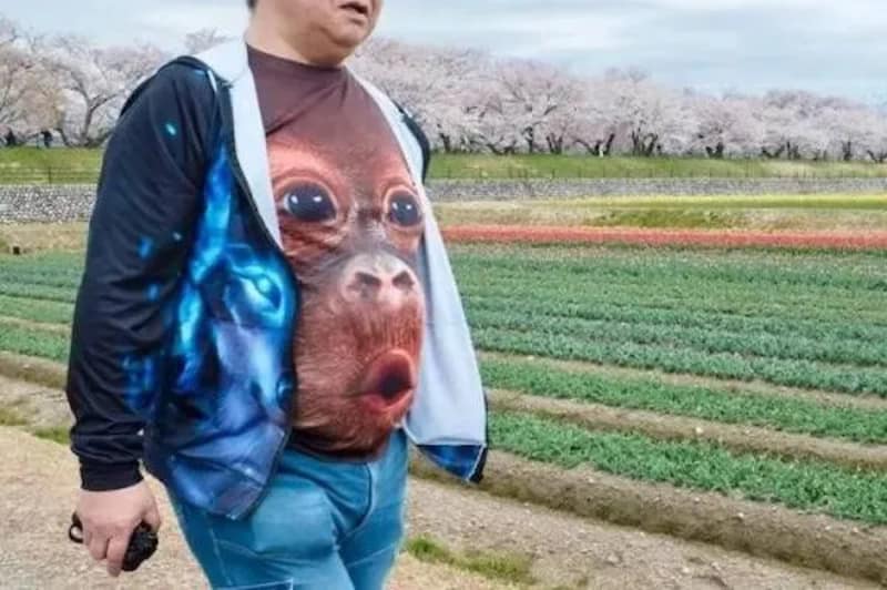 A side-view of a man walking past cherry blossom trees wearing a graphic t-shirt with a giant, wide-eyed baby orangutan face stretched across his stomach, making an excellent addition to this digital meme dump.