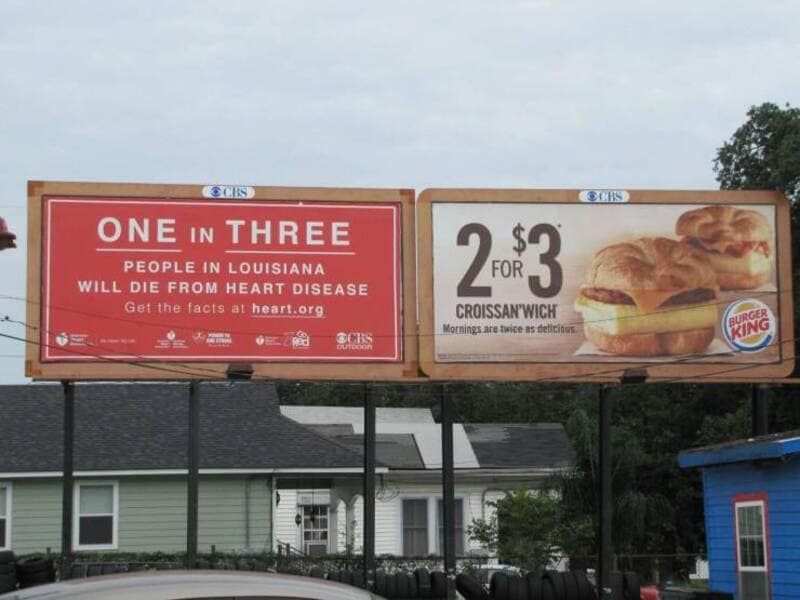 Two side-by-side city billboards create a dark comedy funny meme by placing a grim Louisiana health warning about heart disease directly next to a colorful Burger King advertisement for cheap breakfast Croissan'wiches.