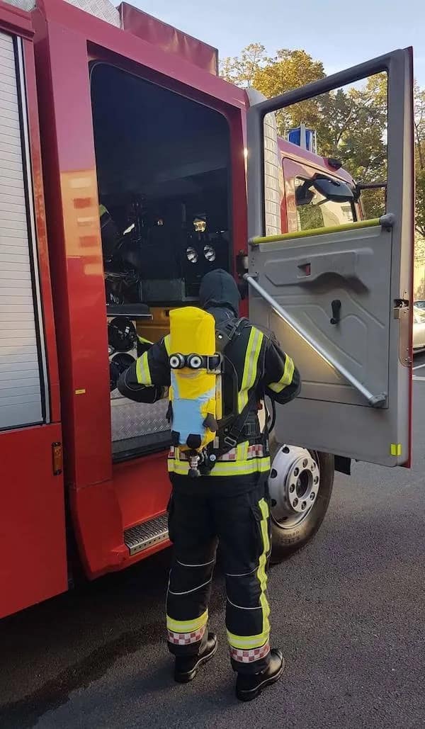A firefighter standing next to an open engine bay door wears their full protective turnout gear, featuring a custom yellow Minion character protective sleeve slipped over their emergency breathing oxygen tank in a funny real-life snapshot.