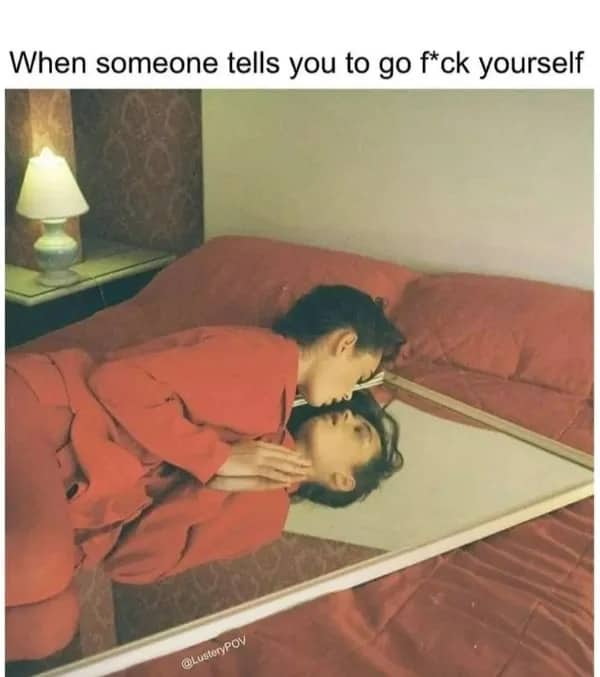 Woman in red suit lying on bed kissing her own reflection in a large mirror.