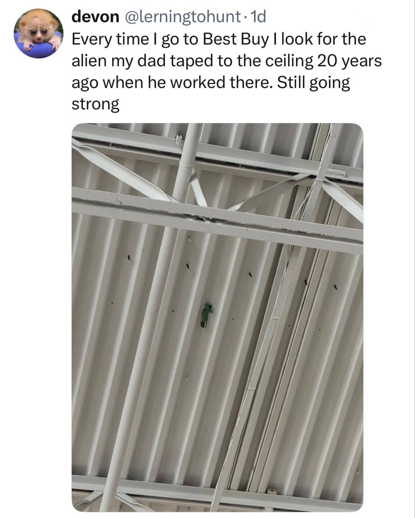 A small green toy alien taped to a white corrugated metal warehouse ceiling.