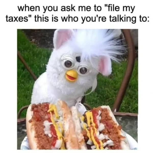 A white Furby toy sitting behind three chili dogs with a caption about being incapable of filing taxes.
