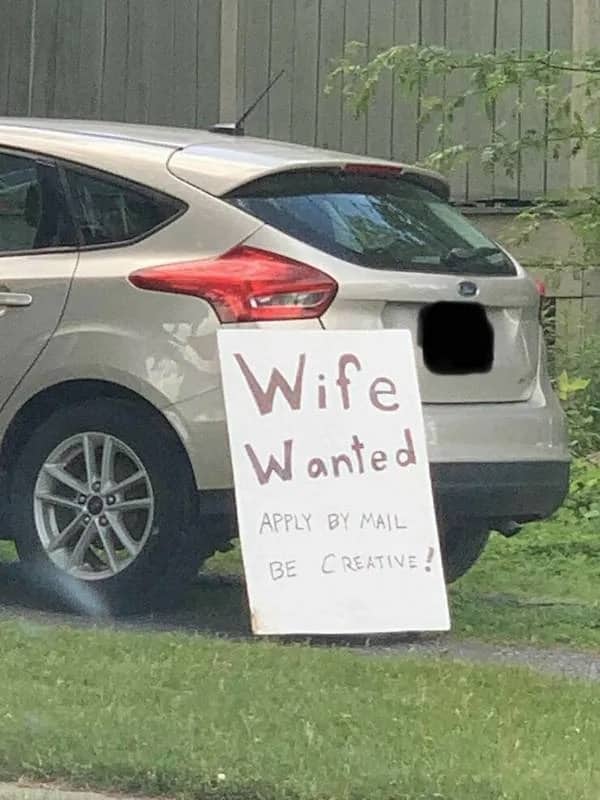 A handwritten sign saying Wife Wanted Apply By Mail leaning against the back of a car.