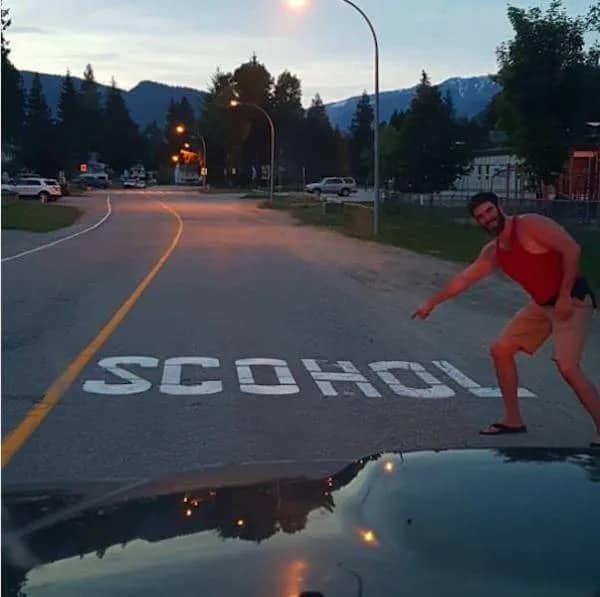 Man pointing at a road marking that has been misspelled as SCOHOL instead of SCHOOL.