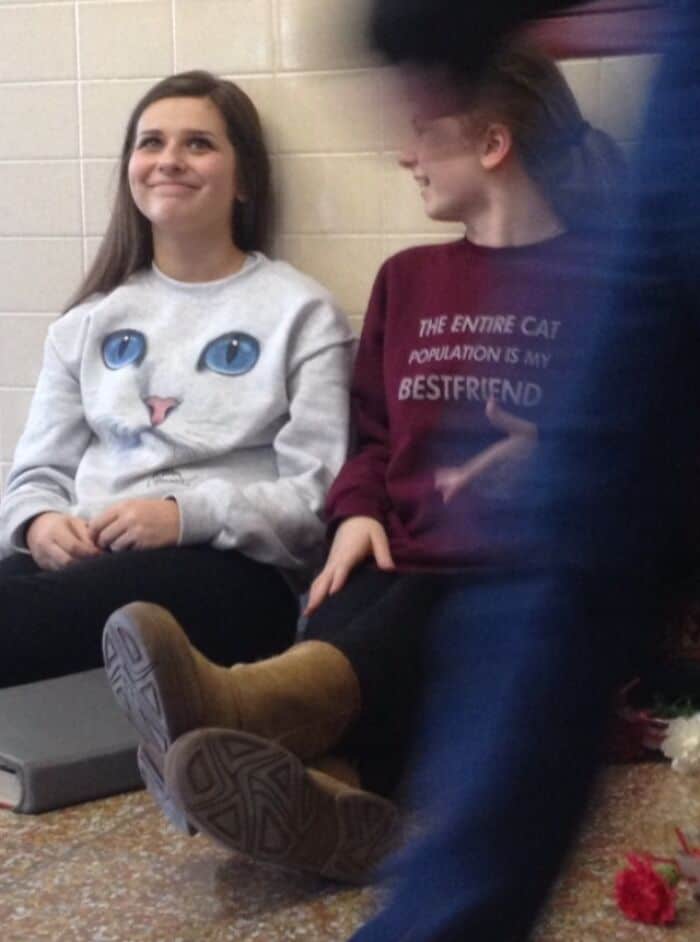 Two girls sit against a wall, one wearing a cat face shirt and the other a cat-themed sweatshirt.