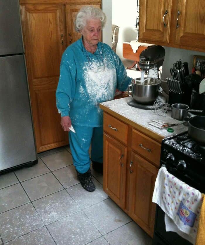 An elderly woman stands in her kitchen completely covered in white flour after a baking mishap.