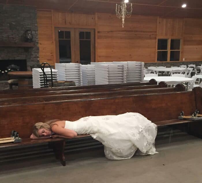 A bride in a white wedding gown lies facedown on a wooden church pew, exhausted.