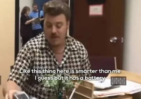 Ricky looks confused in an office with other people blurred in the background.