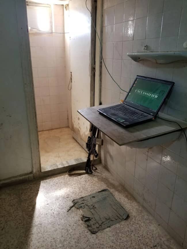 A laptop setup on a makeshift wooden shelf inside a dingy, tiled bathroom or shower stall.