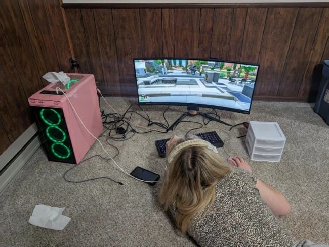 A gamer lying flat on a carpeted floor playing on a curved monitor with a pink PC case nearby.