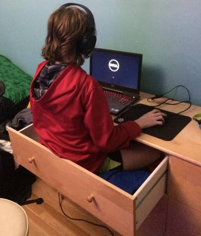A person sitting inside a pulled-out wooden desk drawer while using a laptop on the desk surface.
