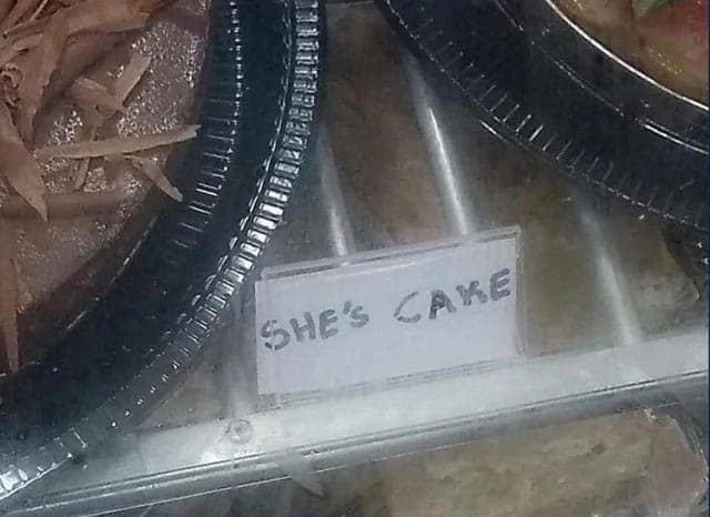 A bakery display sign featuring a spelling fail where a cake is labeled as "SHE'S CAKE" instead of "Cheesecake."