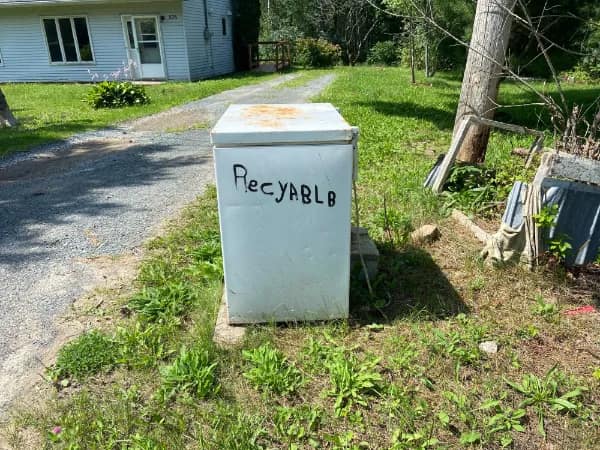 An old, weathered white chest freezer sitting out on a grassy lawn functions as a funny english fail with "RECYABLB" crudely hand-written in black marker across the front panel.