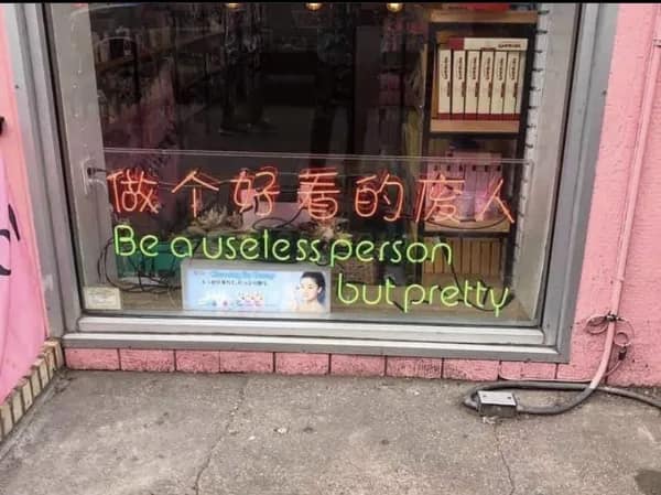 A glowing green neon sign inside a pink storefront window showcases an iconic, strangely motivational translation fail that reads "Be a useless person but pretty" below the original Chinese characters.