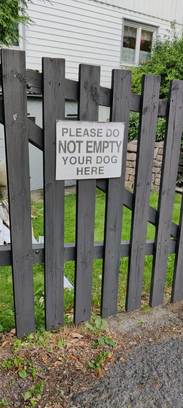 A white metal notice pinned to a dark wooden picket fence features an awkward, word-swapped english fail politely stating "PLEASE DO NOT EMPTY YOUR DOG HERE."