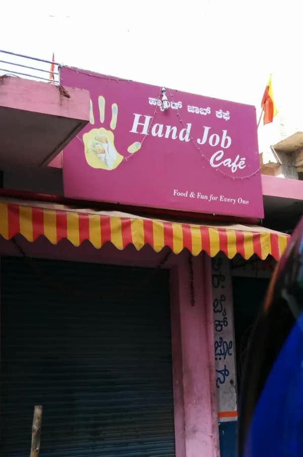 A bright pink commercial storefront sign features a hilarious cross-language translation fail by naming the establishment the "Hand Job Cafe" above the tagline, "Food & Fun for Every One."