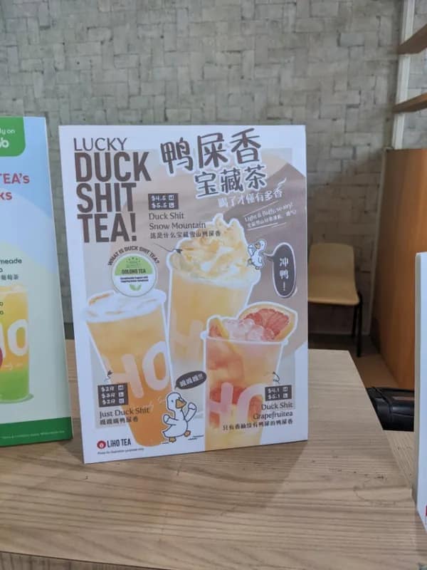 A promotional retail poster at a beverage counter showcases a shocking literal translation fail labeled "LUCKY DUCK SH*T TEA!" alongside a cartoon duck drawing.