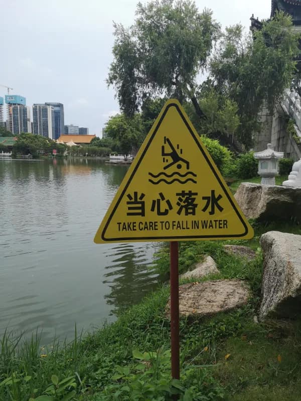 A yellow triangular park caution sign by a lake features a catastrophic translation fail that instructs visitors to "TAKE CARE TO FALL IN WATER" beneath an icon of an upside-down tumbling person.