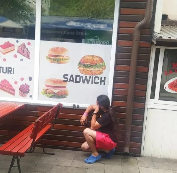 A person crouches in secondary embarrassment on a sidewalk next to a restaurant window display that features a prominent print layout translation fail labeling a cheeseburger as a "SADWICH."