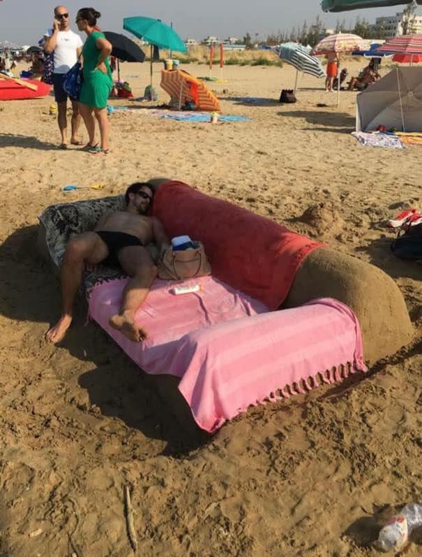A humorous vacation meme showing a man relaxing on a full-size sofa custom-sculpted entirely out of beach sand and draped with towels to perfectly mimic a real living room couch setup.