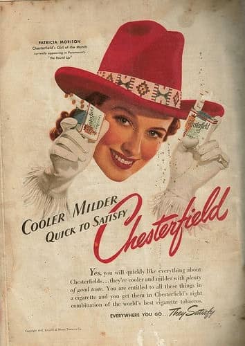 A faded vintage cigarette ad for Chesterfield featuring actress Patricia Morison wearing a wide-brimmed red hat and white gloves. She holds a cigarette pack in each hand near her face, framed by the text "Cooler Milder Quick to Satisfy."