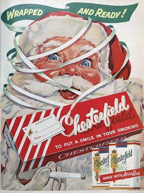 A vibrant vintage cigarette ad for Chesterfield featuring Santa Claus tangled in green and white festive ribbons. He holds a lit cigarette in one hand and a carton of cigarettes in the other, under the playful slogan "Wrapped AND Ready!"