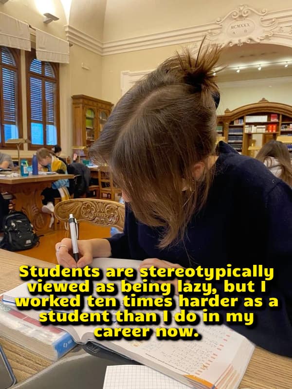A university student with a messy bun focuses intensely on open textbooks at a wooden library desk, serving as the setting for a relatable visual funny shower thought comparing intense school workloads to adult careers.