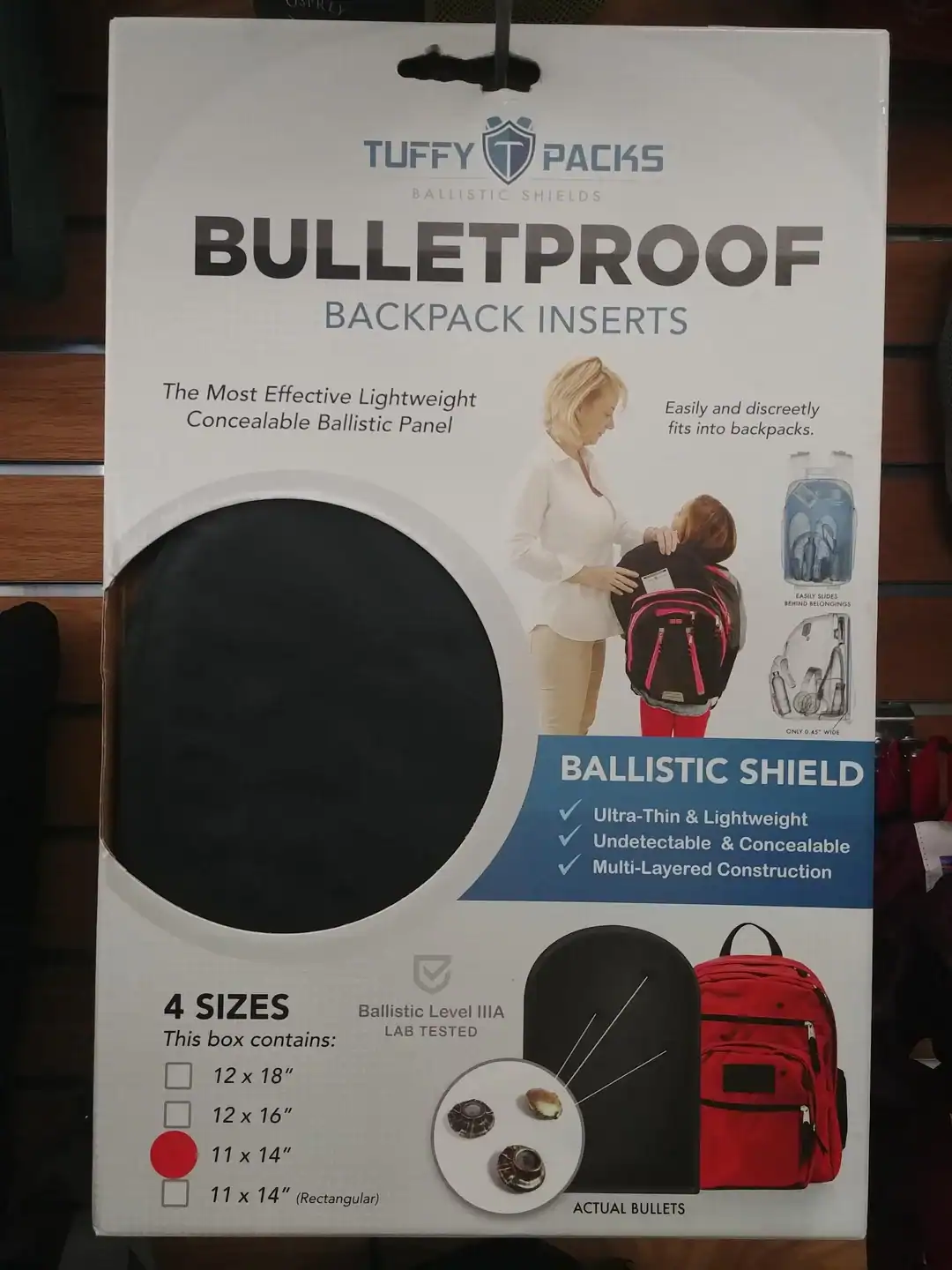 A sobering weird product on a store hanger: "Tuffy Packs" bulletproof backpack inserts, featuring a photo of a mother sliding a ballistic shield into a young girl's pink school bag.