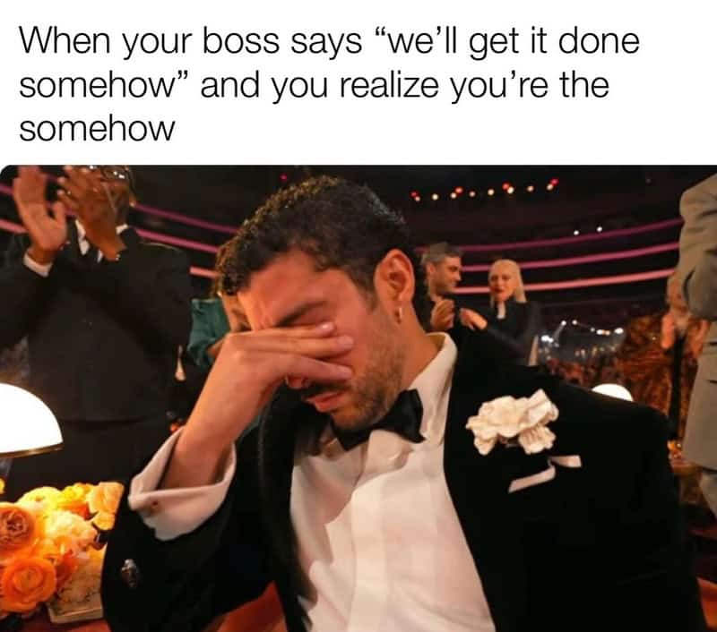 A stressful work meme using a photo of musician Bad Bunny at an awards show. He is wearing a tuxedo and has his hand over his eyes in a gesture of absolute defeat. The text hits home, describing the moment a boss says a project will be finished "somehow," and you realize you are the "somehow."
