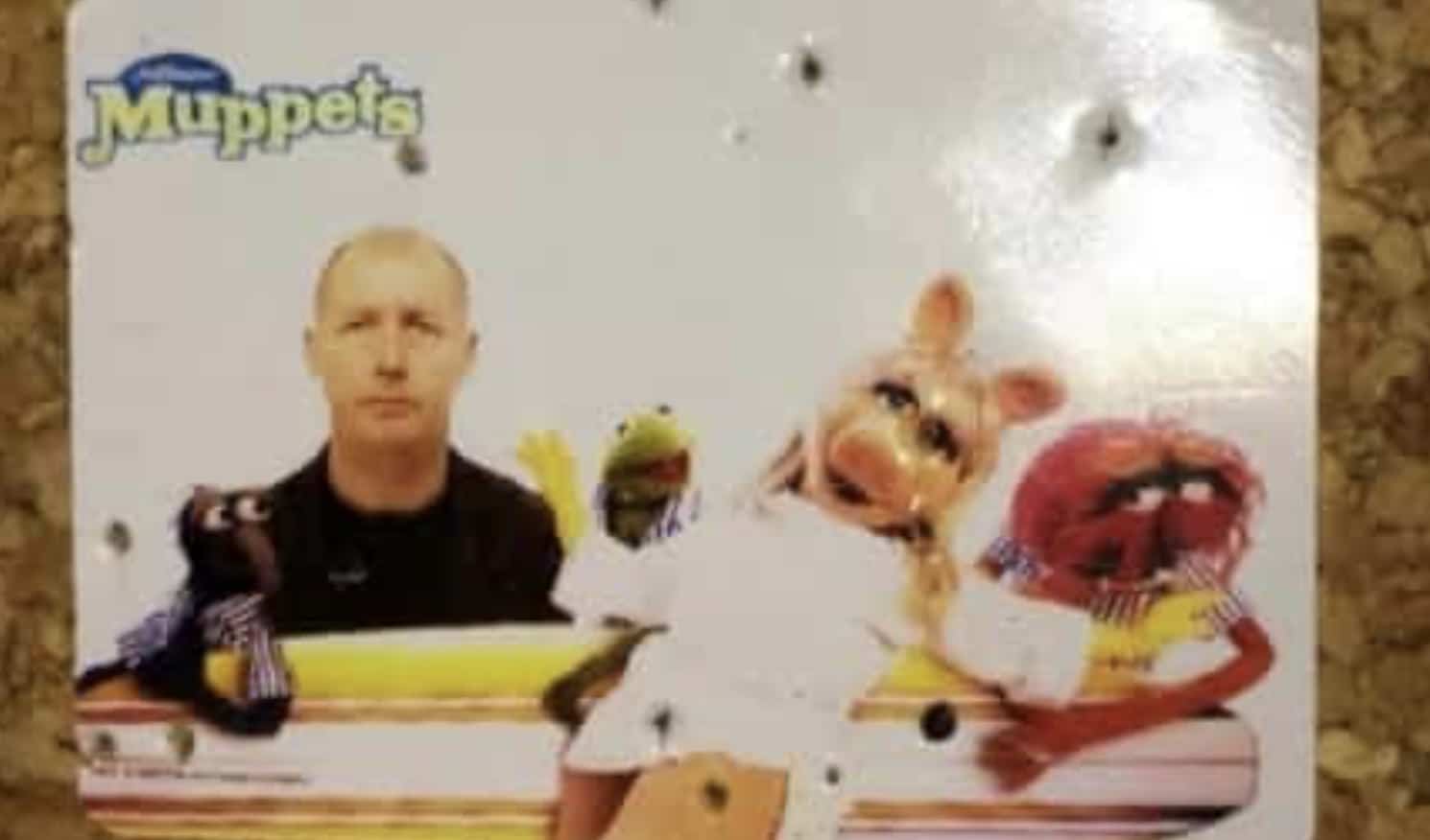 Bald man standing solemnly next to Kermit, Miss Piggy, and Animal Muppets on a surfboard.