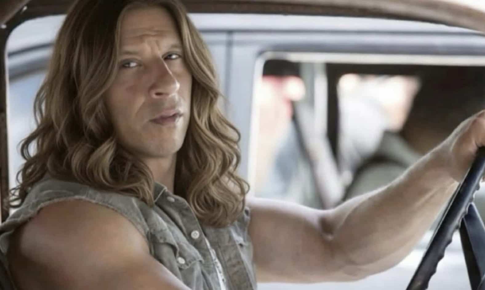 Vin Diesel edited with long, wavy blonde hair sitting behind the wheel of a car.