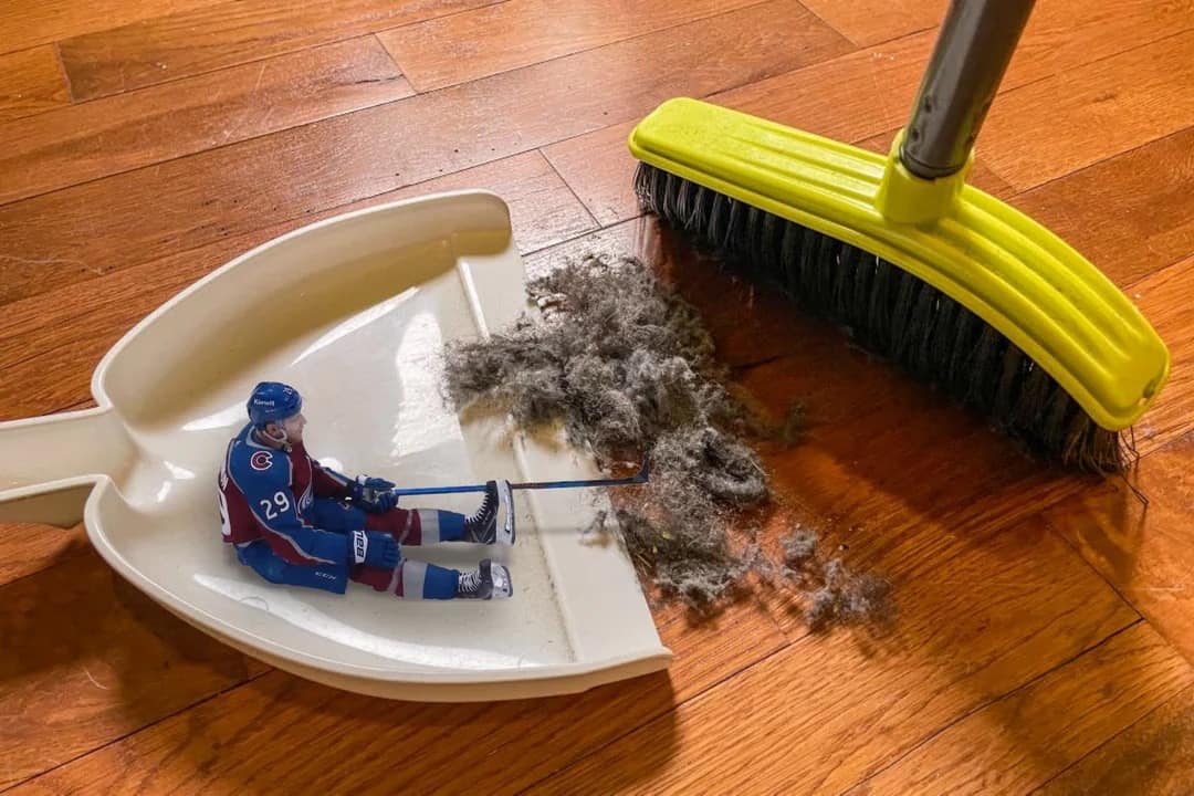 A literal "sweep" photo shared as an elite 2026 Stanley Cup Finals meme shows a yellow kitchen push-broom sweeping a massive pile of grey dust bunnies and a miniature Nathan MacKinnon toy action figure across a hardwood floor into a white plastic dustpan.