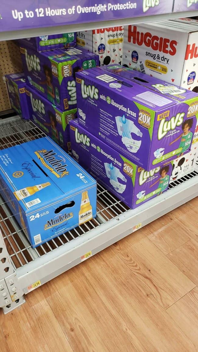 A store shelf places a case of Modelo beer directly beside boxes of Luvs and Huggies diapers, creating an accidental adulthood meme about the two core survival supplies of grown-up life: crisis management and overnight protection.