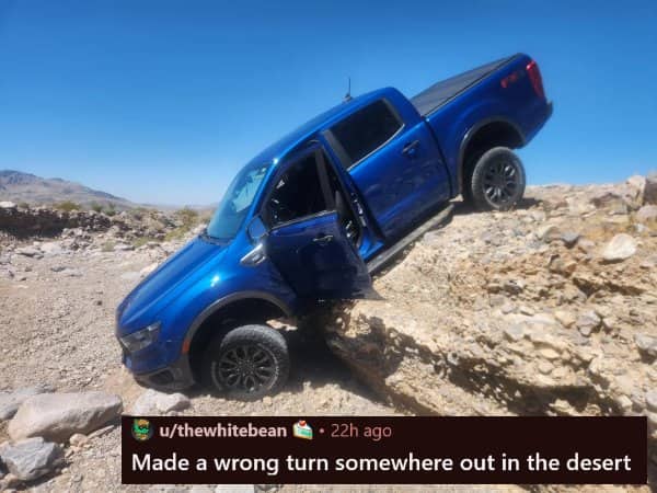 A blue pickup truck high-centered and stuck precariously on a large boulder in the desert.