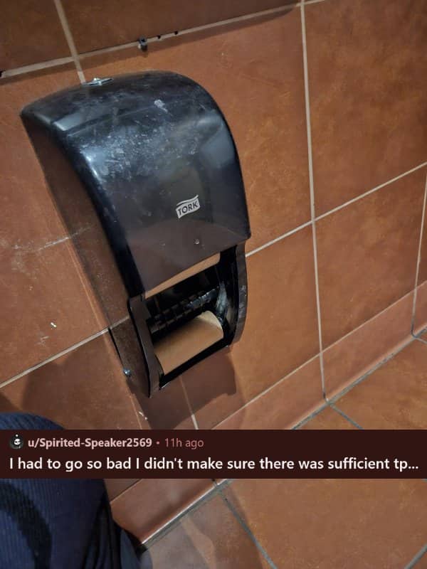 An empty black plastic toilet paper dispenser on a brown tiled wall showing only a cardboard roll.